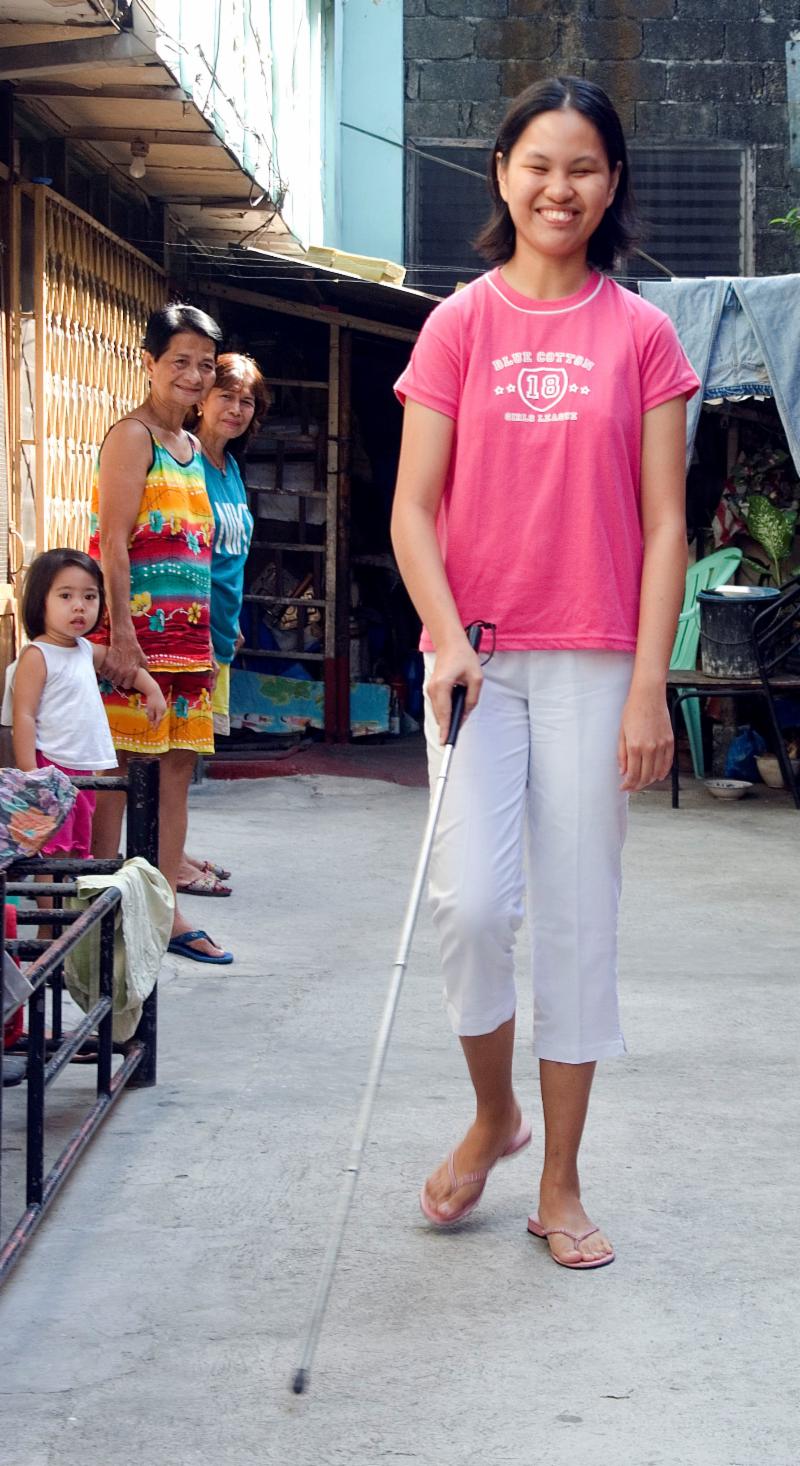 Smiling Filipino young woman walks with cane as others watch with interest